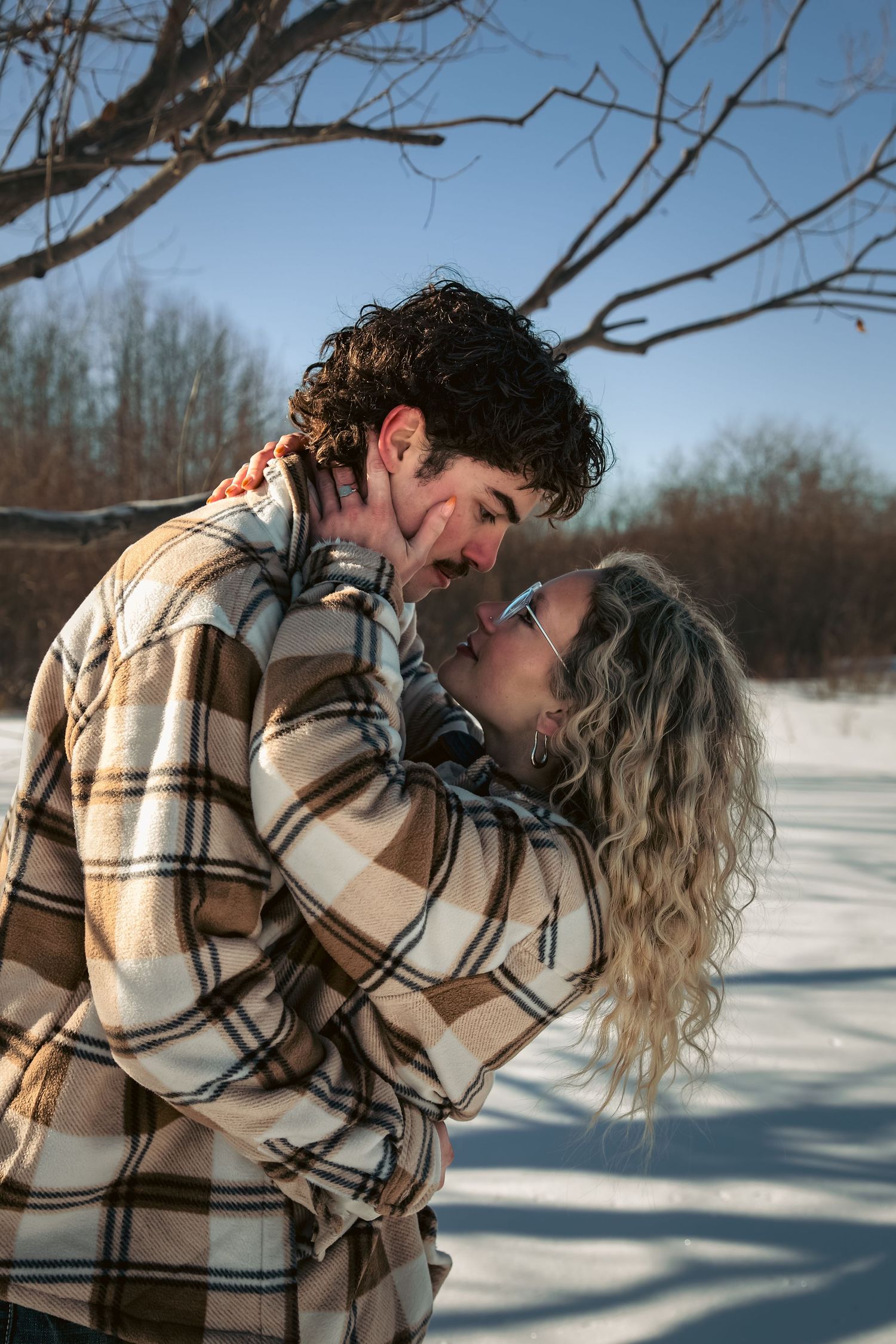 Couple in matching plaid jackets laughing and almost kissing in a sunny snowy field.