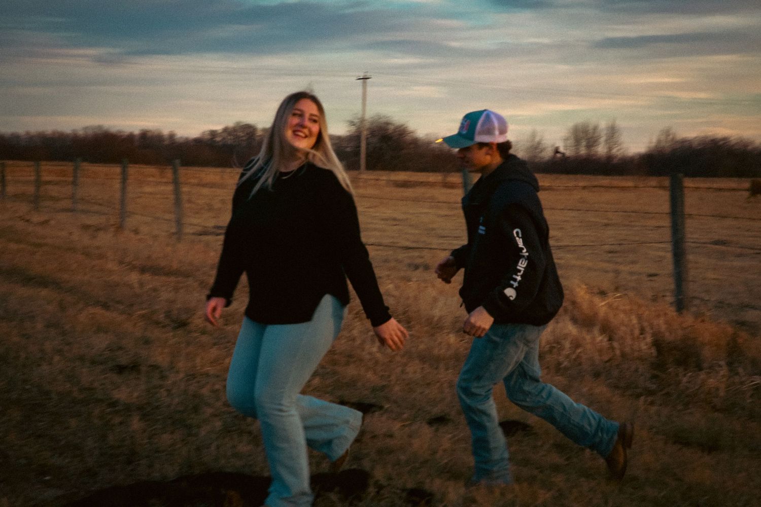 A laughing couple walks hand in hand along a fence line in a golden-hour rural field setting.