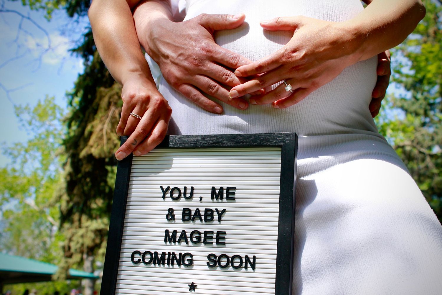 Couple forming a heart shape over a pregnant belly beside a Baby Magee Coming Soon letterboard sign.