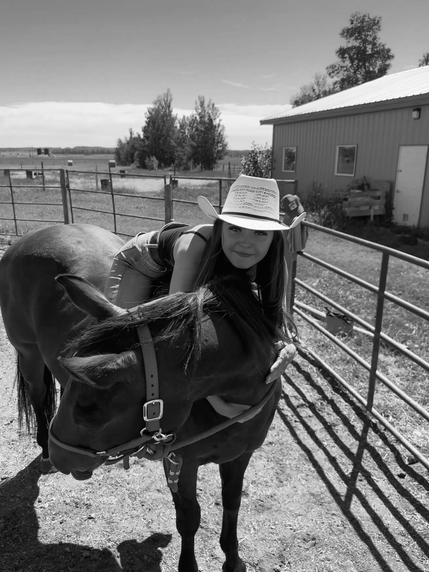 A woman in a cowboy hat stands beside a saddled horse in a ranch corral, black and white photo.