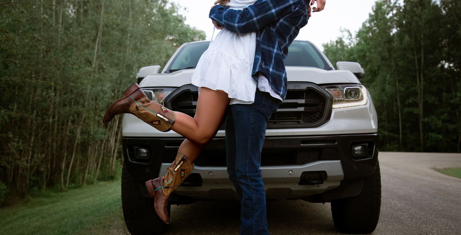 Couple sharing a romantic embrace leaning against the hood of a white truck on a wooded road.