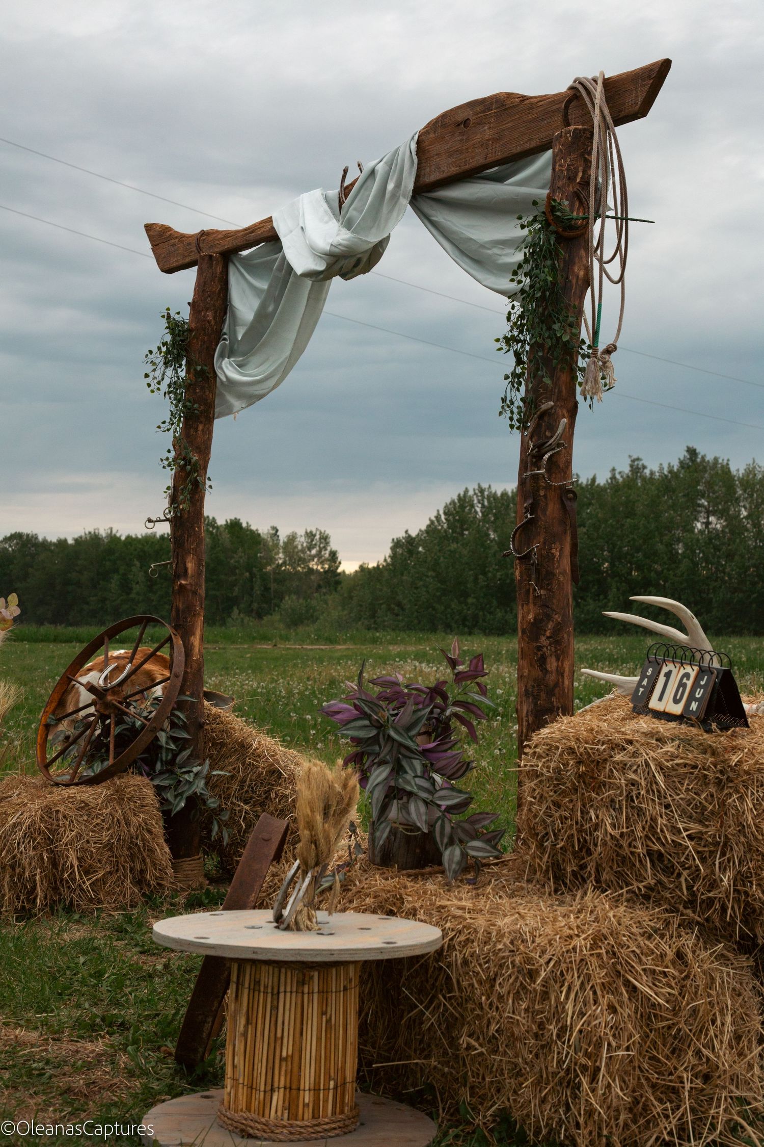 Rustic country wedding arch with wooden posts, white fabric draping, hay bales, purple plants, and wagon wheel.