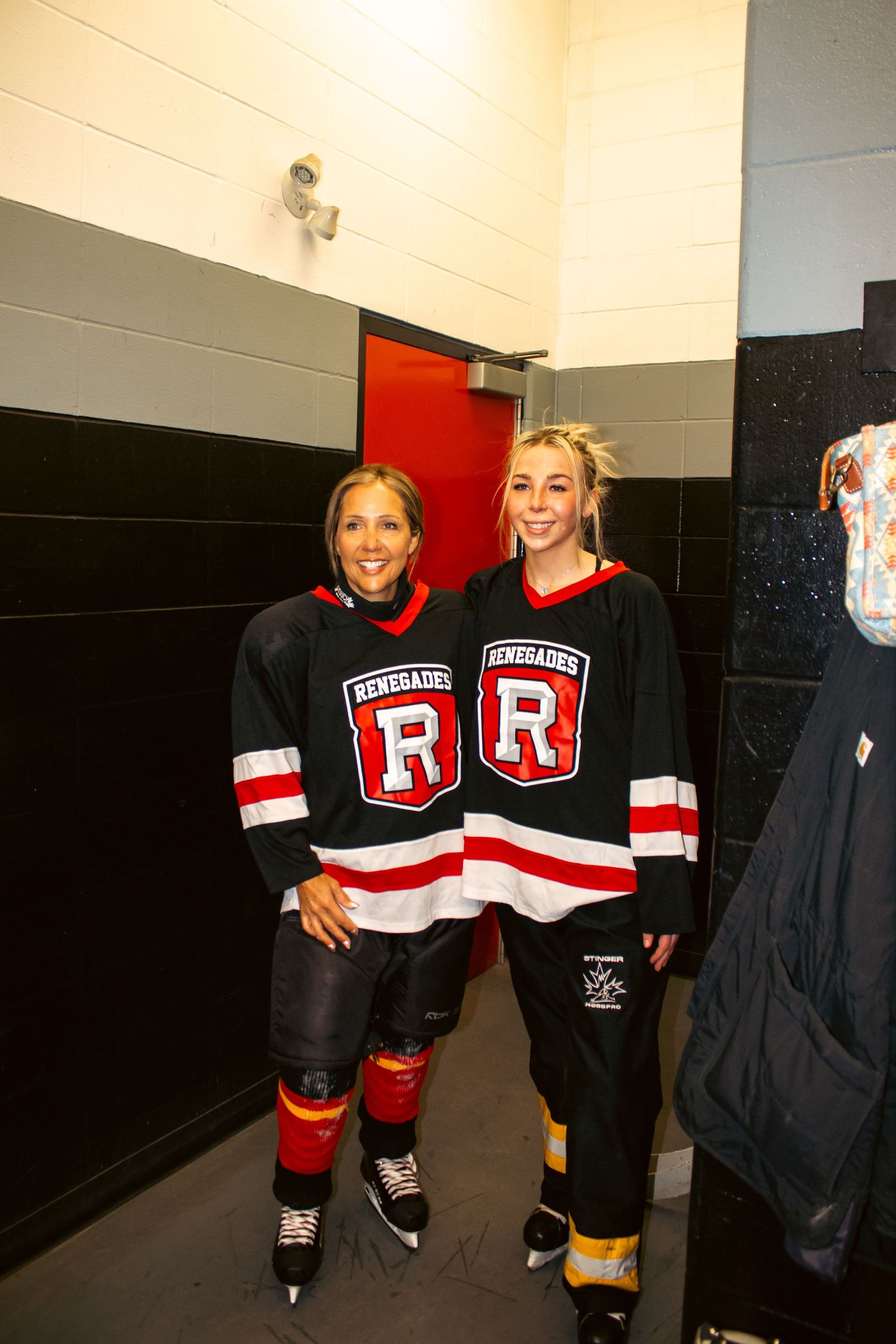 Two women smiling in Renegades hockey jerseys standing in arena locker room hallway.