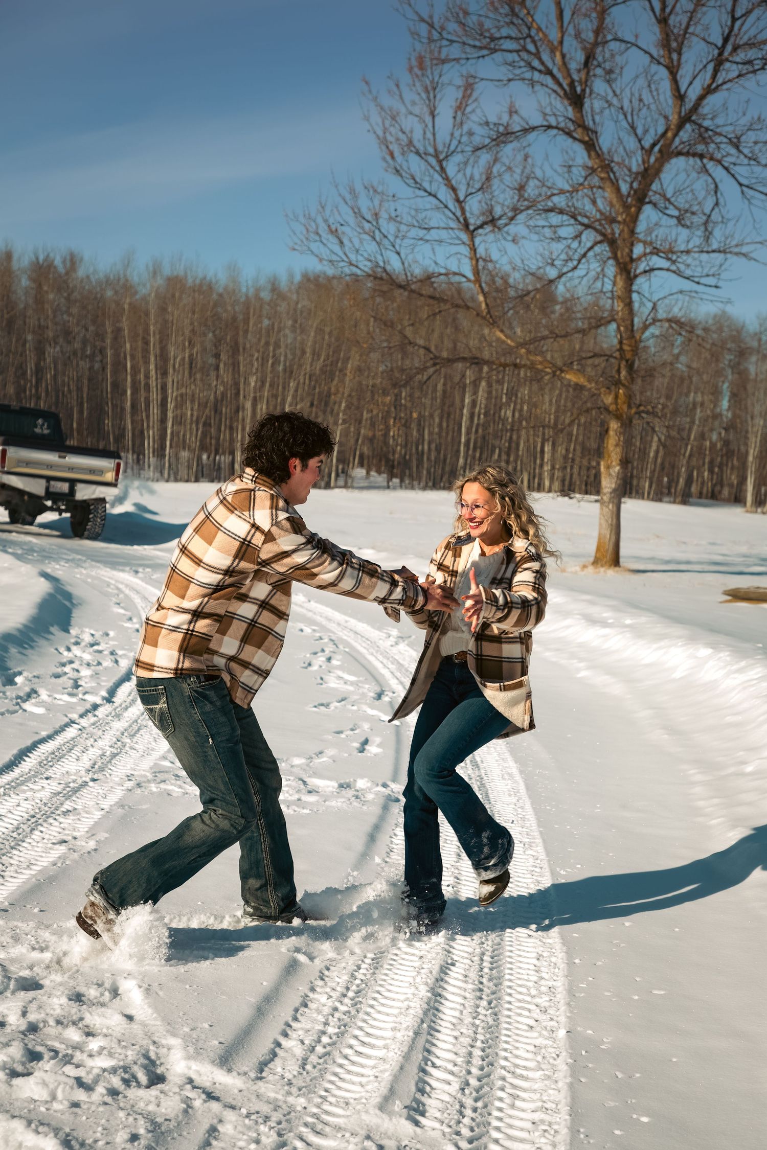Playful couple in matching plaid jackets spinning and laughing together on a snowy country road.