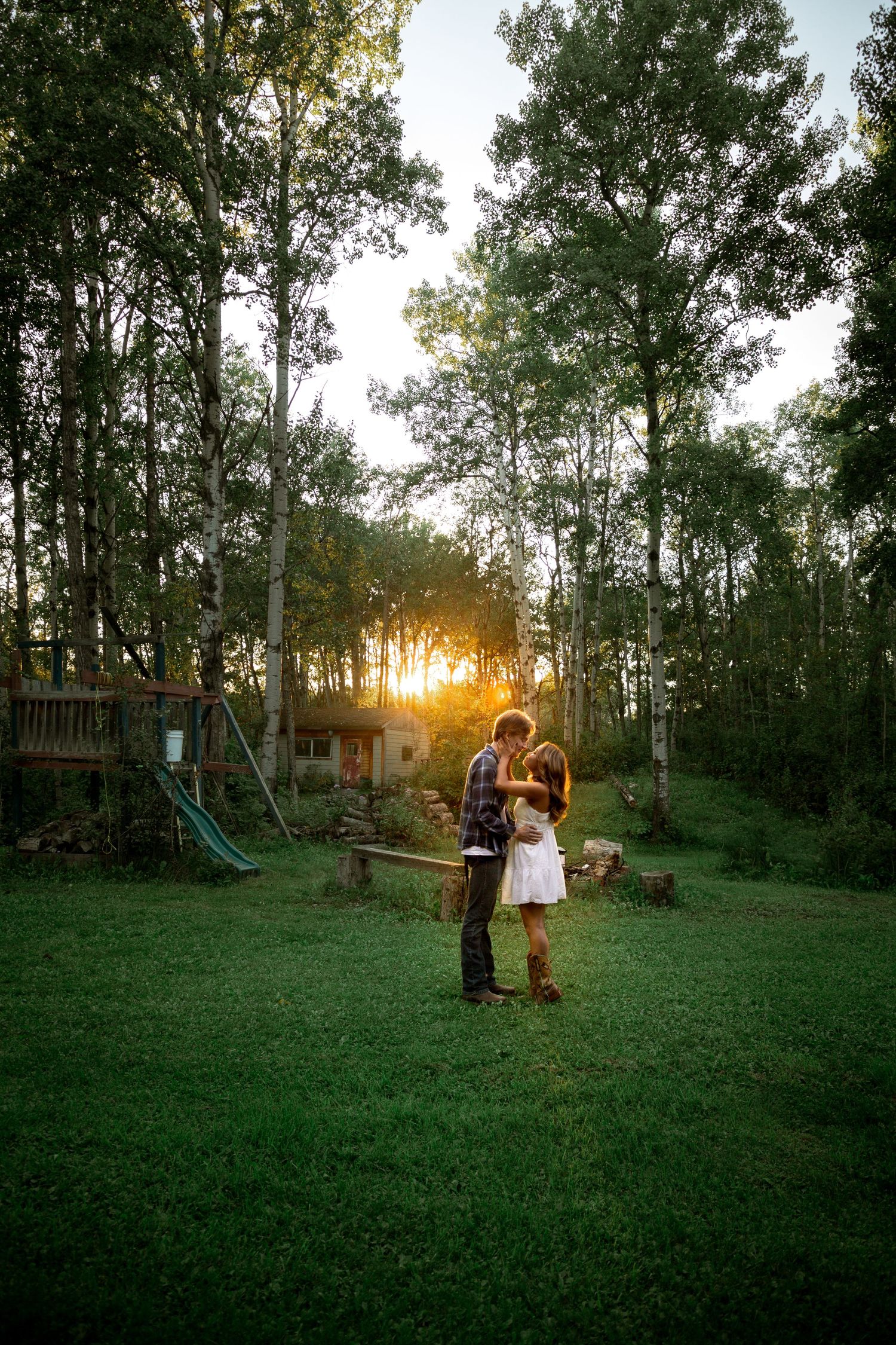 Couple sharing a golden sunset kiss in a lush green backyard surrounded by tall trees.
