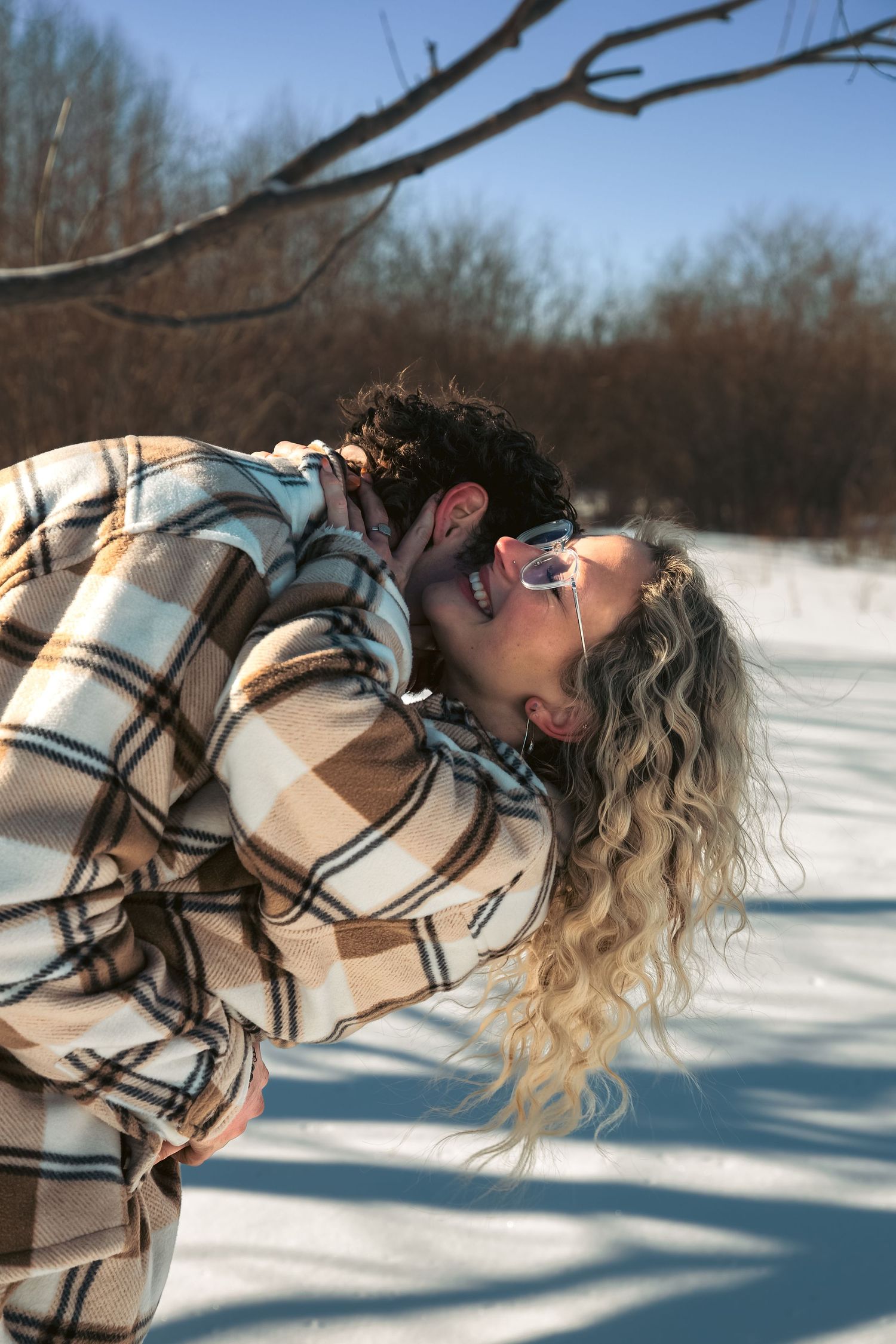 Couple in matching plaid shackets sharing a passionate dip kiss in a snowy winter field.