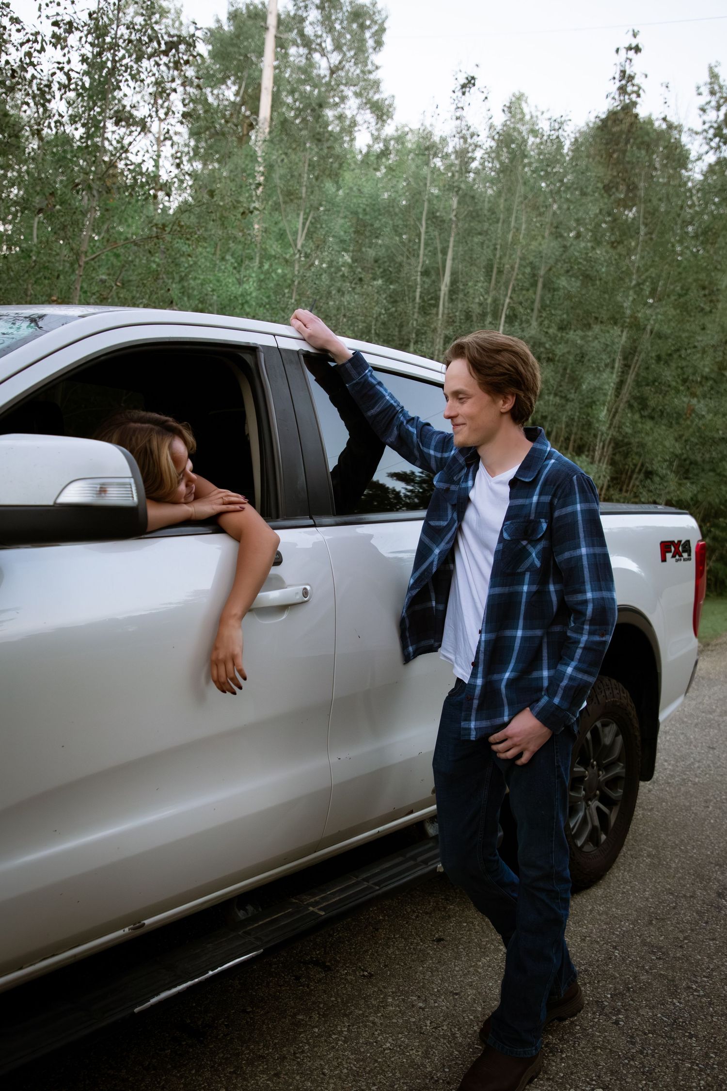 Young man in plaid shirt talking to woman leaning out of white pickup truck window outdoors.