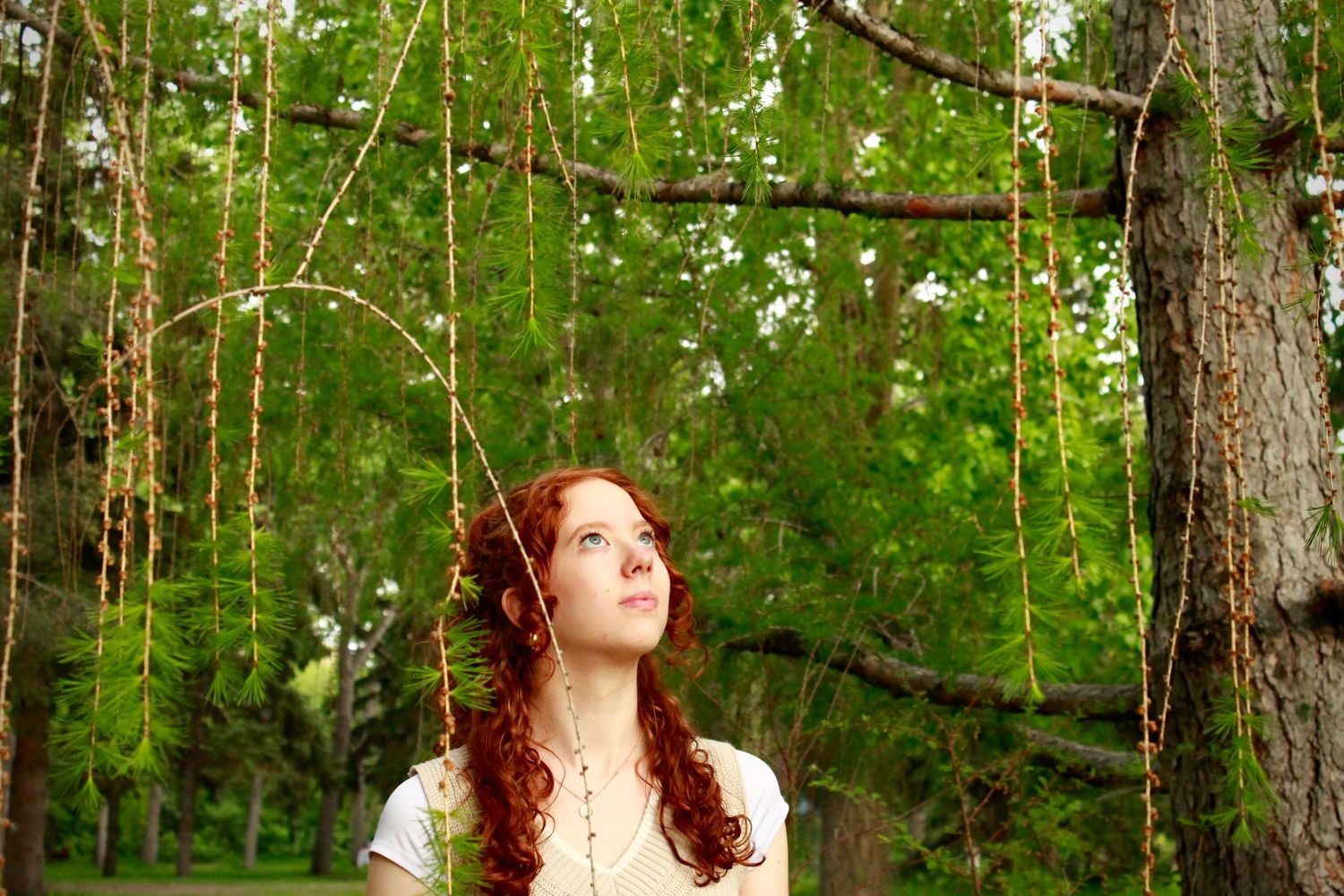 Young redhead woman looking up through hanging green willow branches in a lush summer park setting.