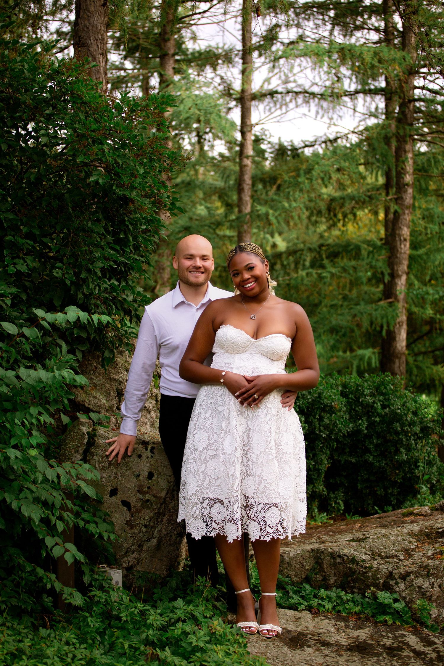 Smiling interracial couple posing together in a lush forest setting, bride wearing a white lace dress.