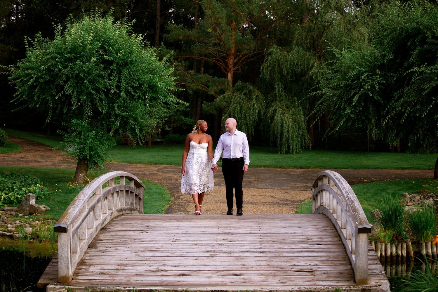 Couple holding hands walking across a wooden bridge in a beautiful green park setting.