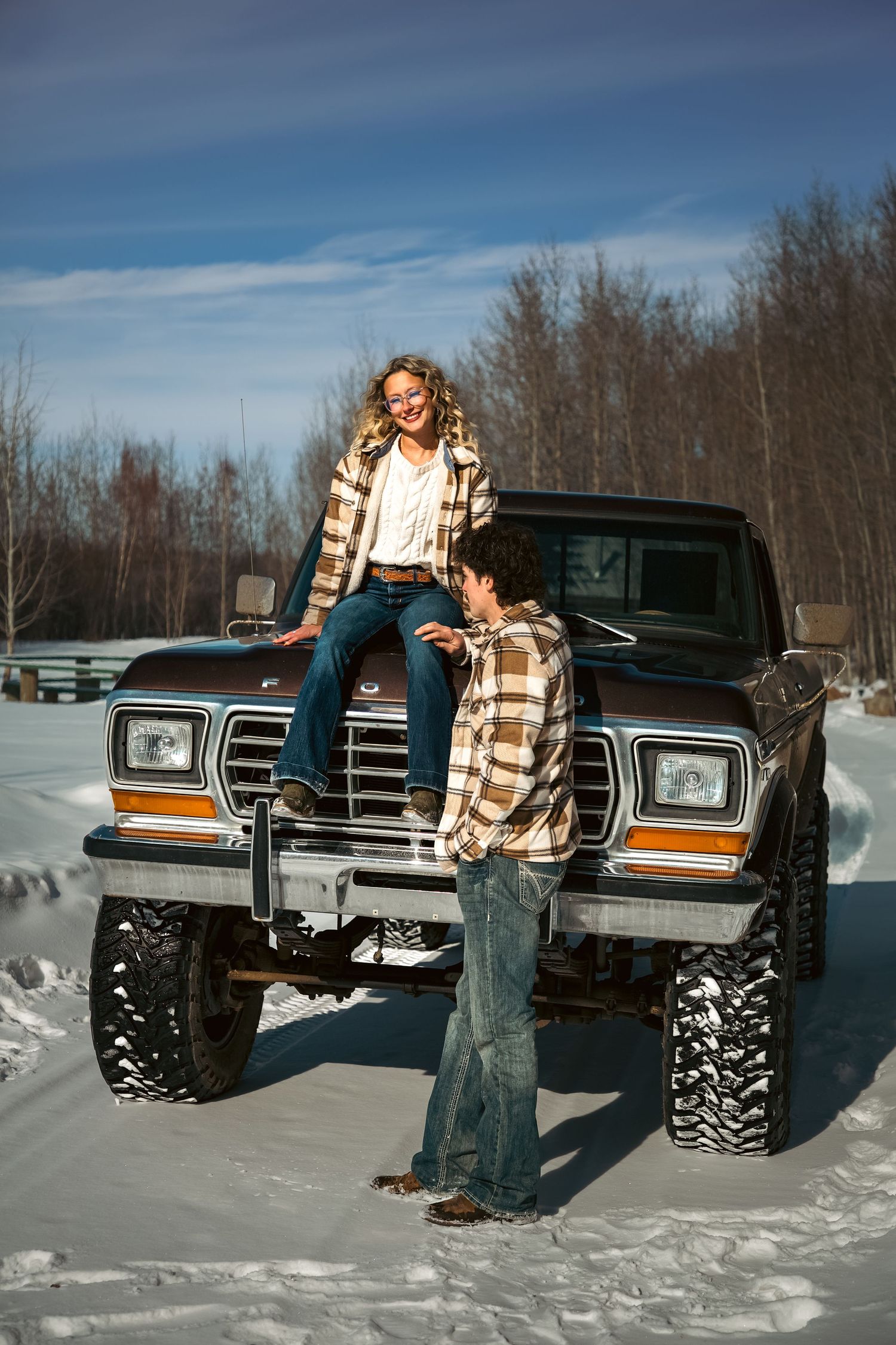Happy couple in matching plaid shirts posing with a vintage Ford pickup truck in winter snow.