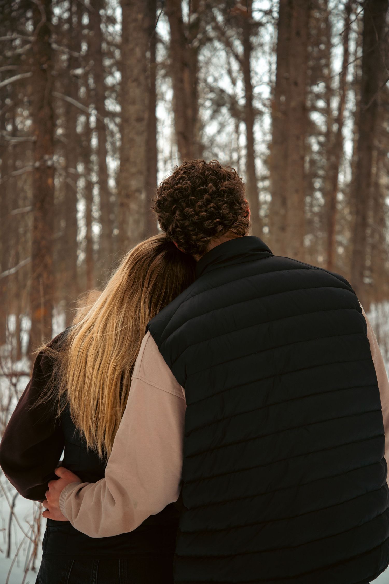 Couple viewed from behind embracing while looking into a snowy pine forest on a winter day.
