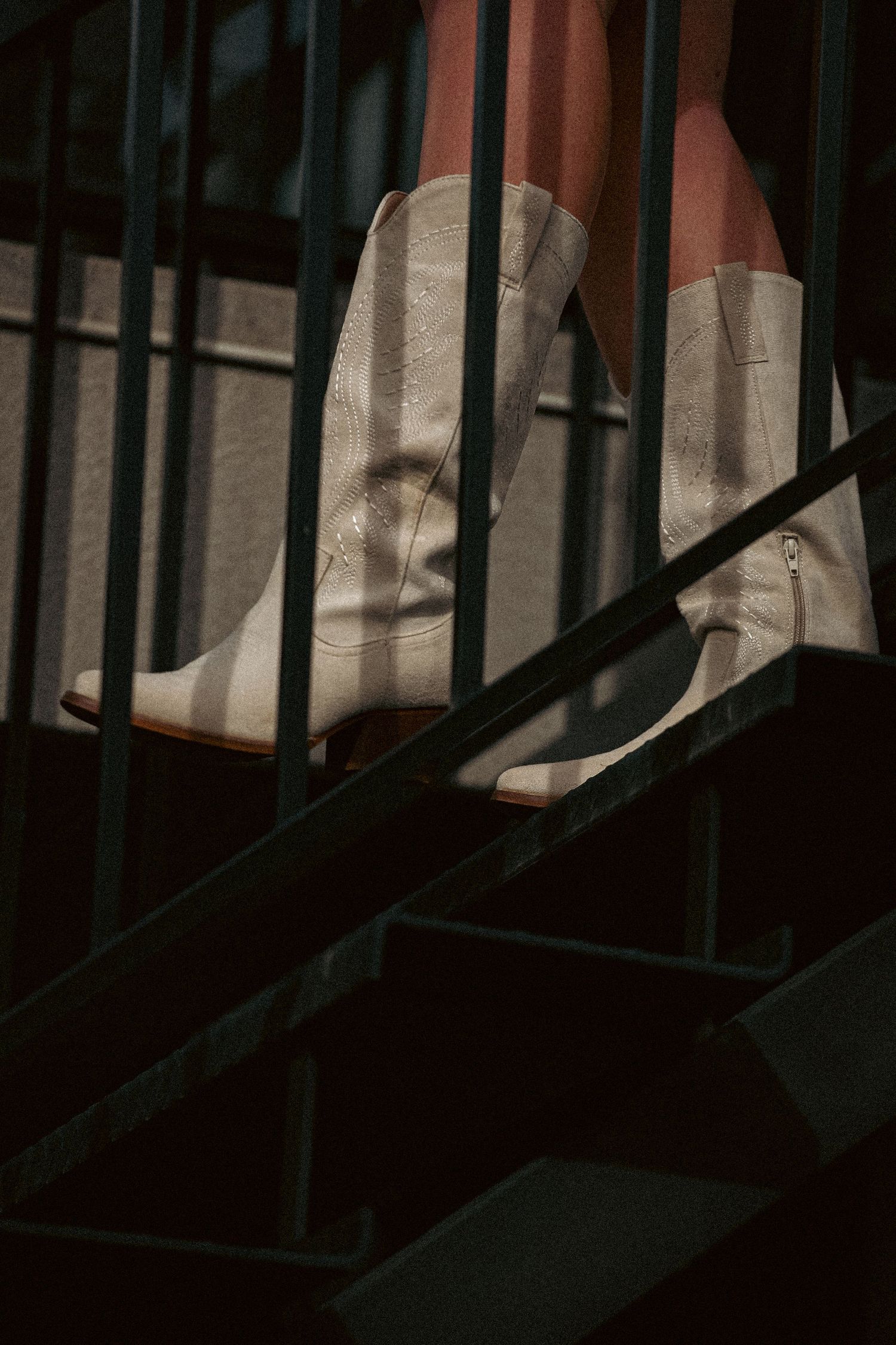 Close-up of white cowboy boots on dark metal outdoor staircase with dramatic shadow lighting.