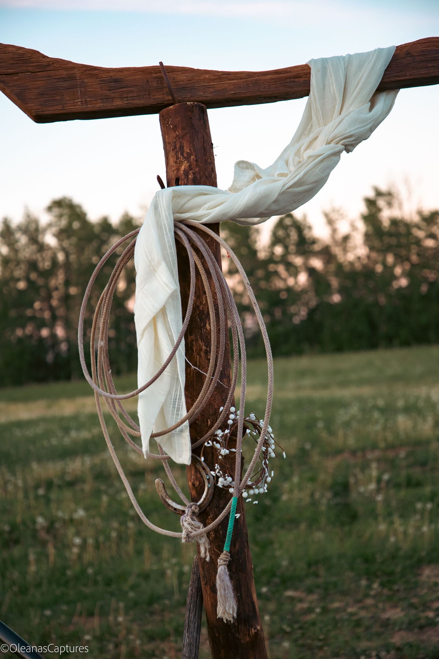 Rustic wooden wedding arch post with coiled lasso rope, white fabric draping, and baby's breath wreath.