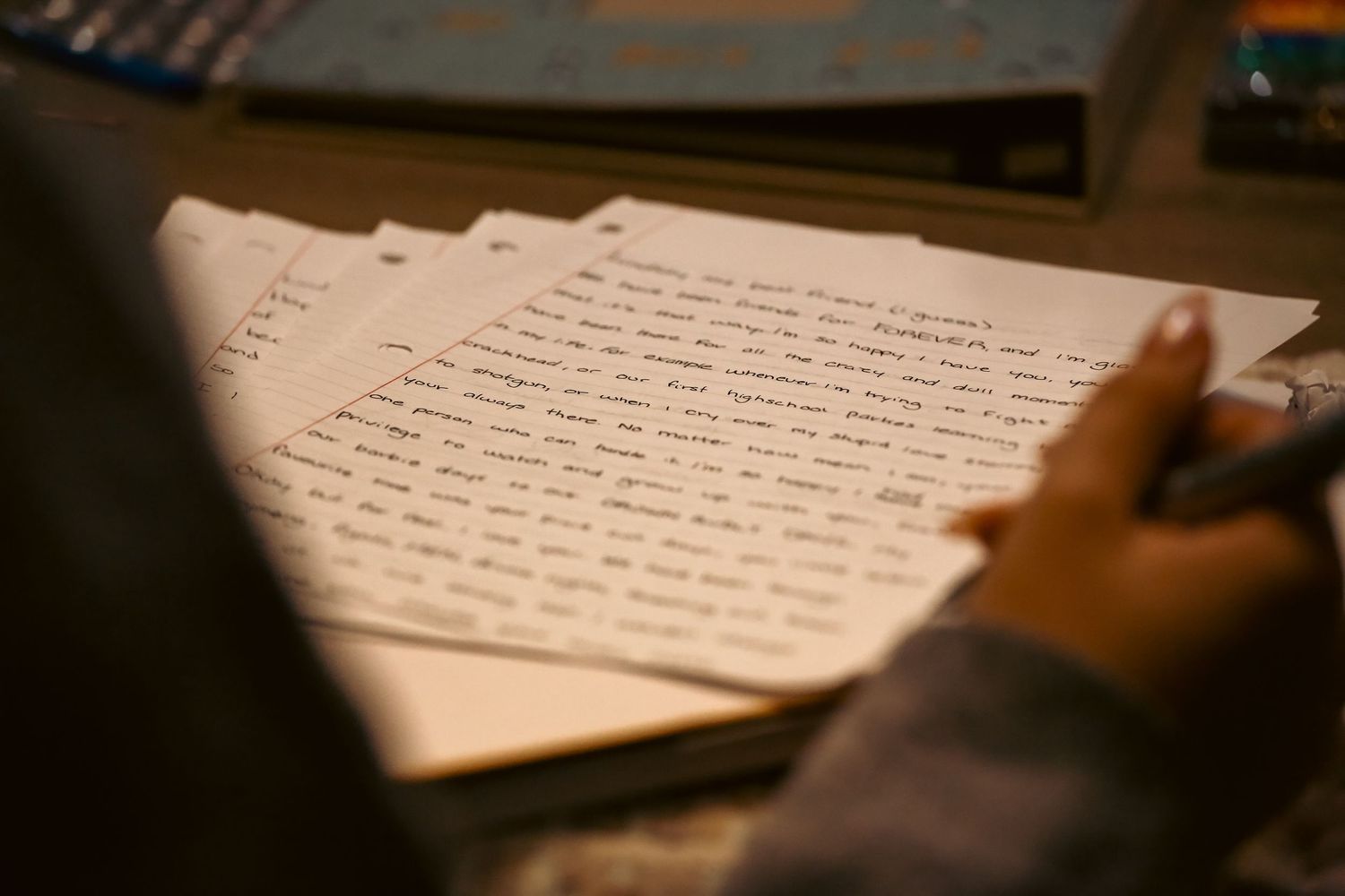 Handwritten pages of cursive writing stacked on a desk in warm ambient lighting.