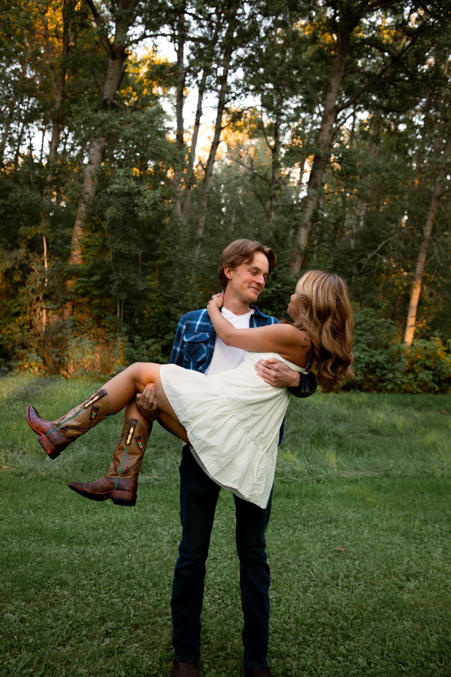 Man lifting woman in white dress and cowboy boots in a sunlit green forest clearing.