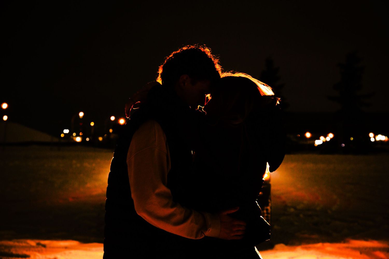 Romantic couple silhouetted against warm glowing lights sharing a kiss outdoors at night in winter.