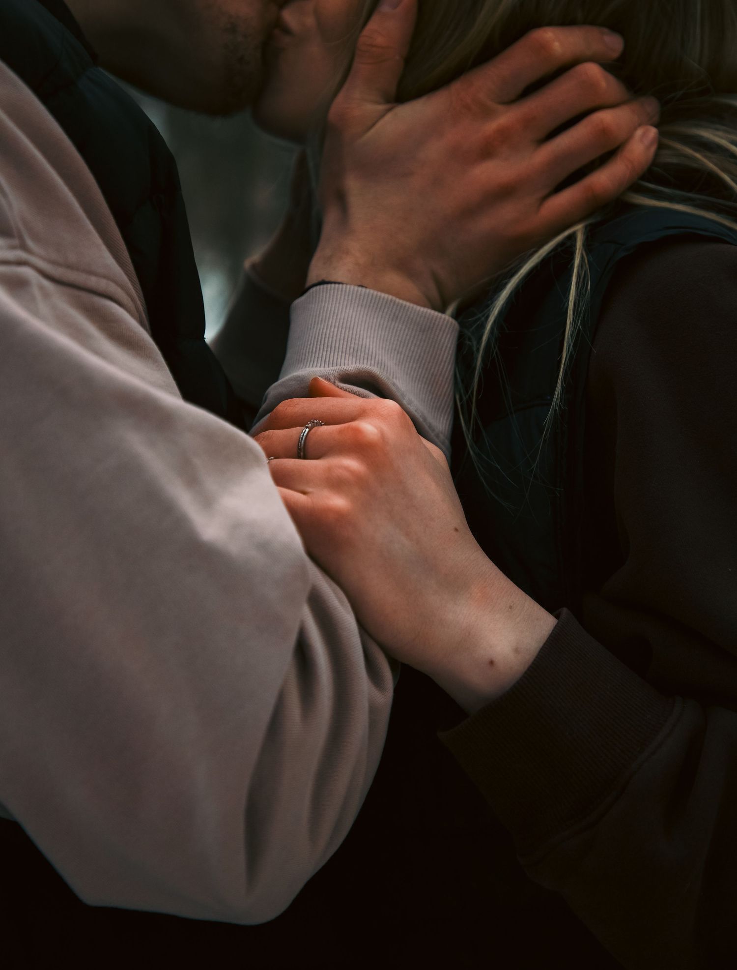 Close-up of couple kissing with focus on hands, one wearing a silver ring, in soft lighting.