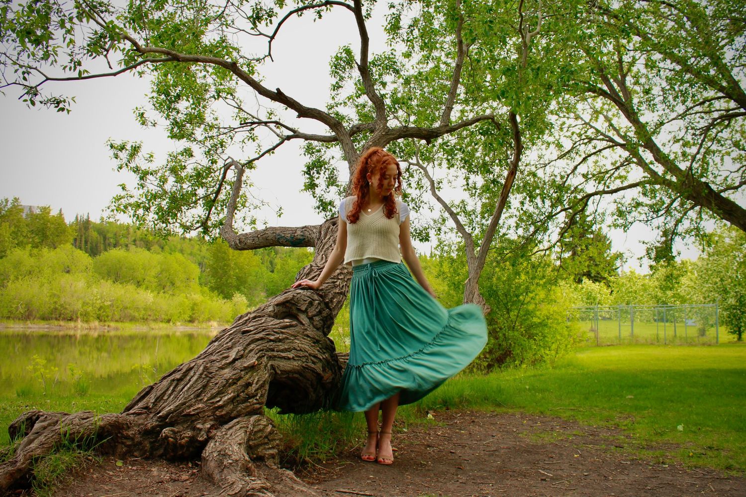 Woman with curly red hair twirls a flowing teal skirt beside a fallen tree trunk in a green park.