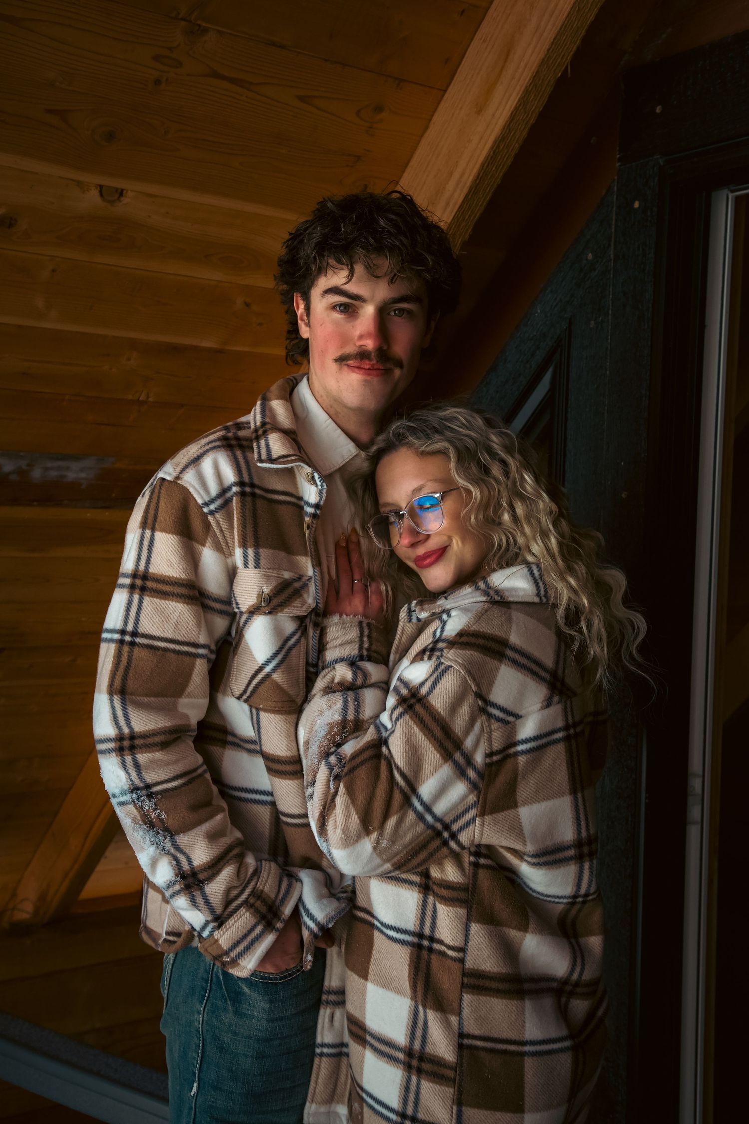 Young couple in matching brown plaid flannel shirts posing together inside a rustic wooden log cabin interior.