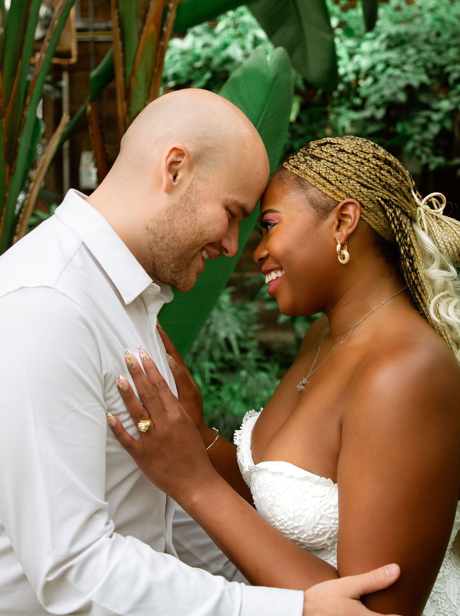 Interracial couple in wedding attire share a romantic forehead touch surrounded by tropical greenery.