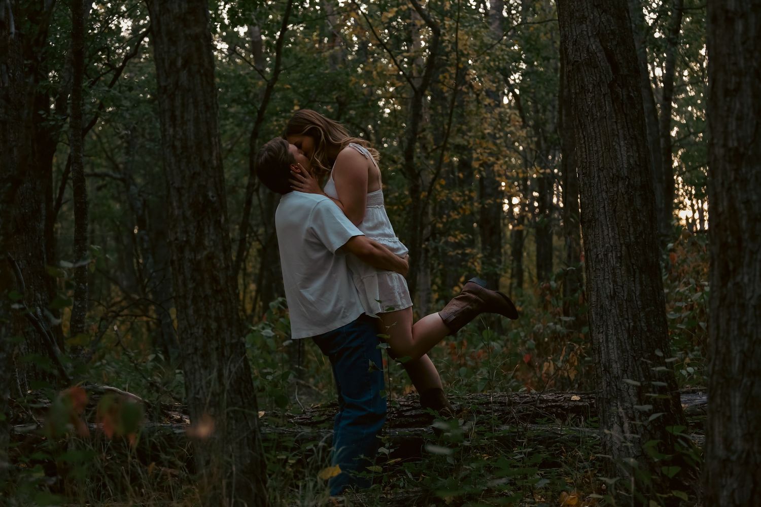 A man lifts and embraces a woman in a white dress among tall trees in a dark forest.