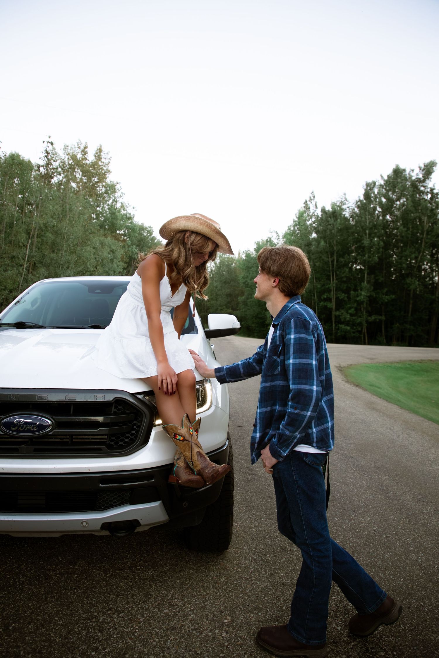 Woman in white dress and cowboy hat sitting on a Ford truck hood with her partner.