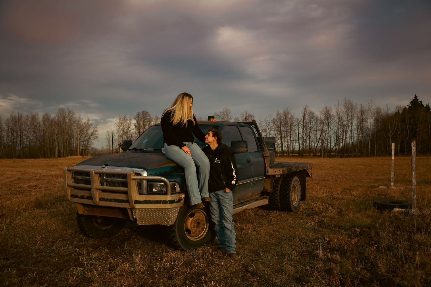 Young couple posing with a vintage truck in a golden-lit open field at dusk.