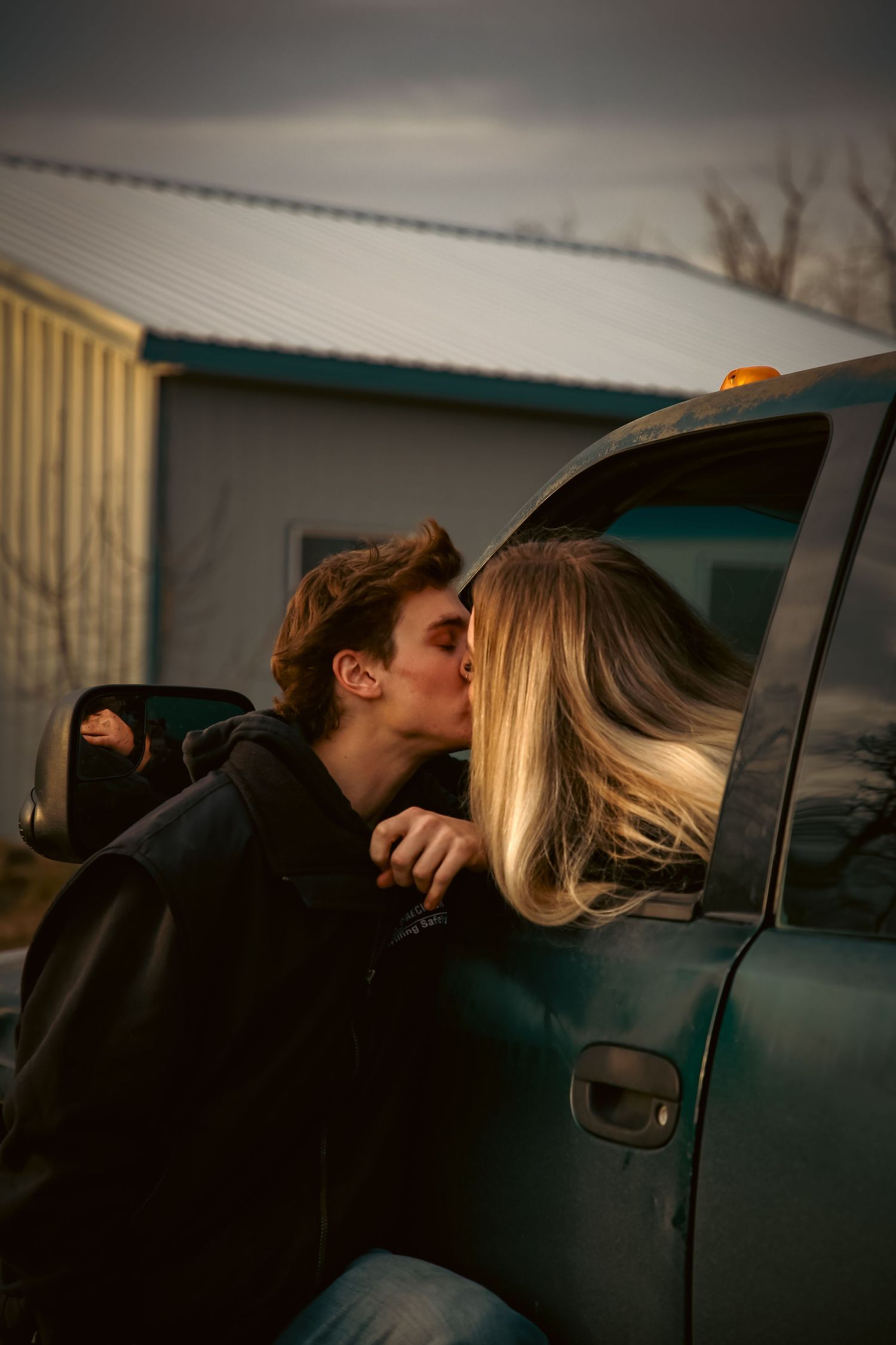 A young couple shares a romantic kiss through a truck window at dusk near a rural building.
