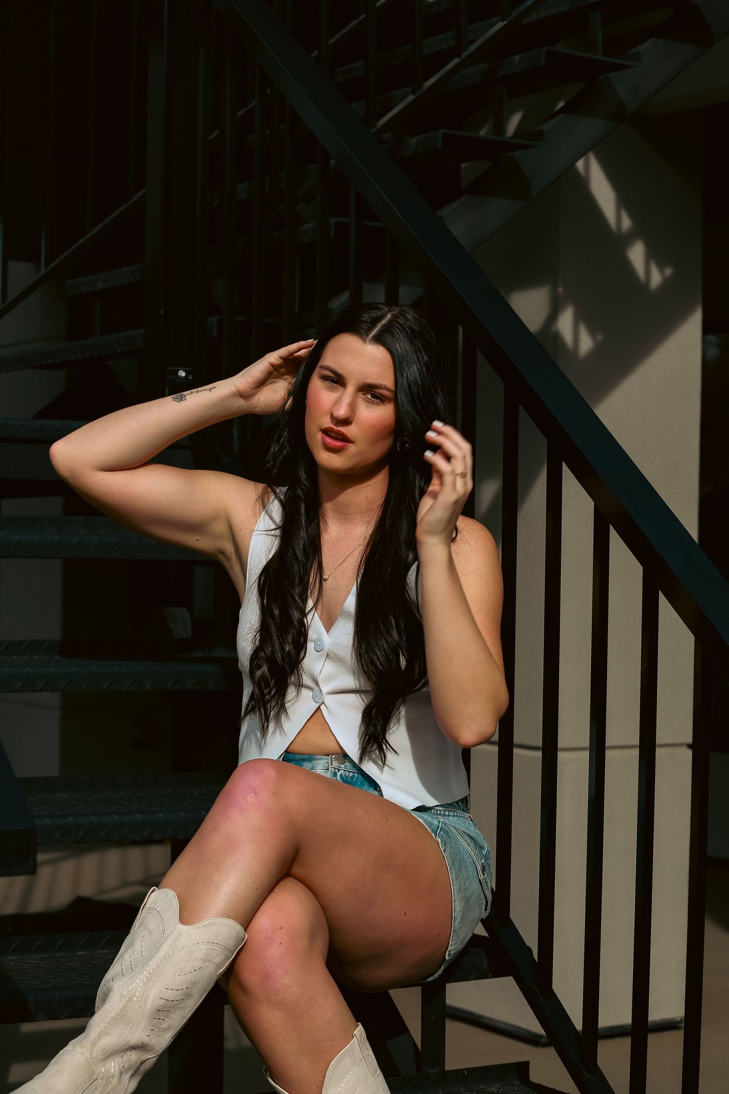 Young brunette woman in white crop top and denim shorts sitting on metal stairs wearing white cowboy boots.