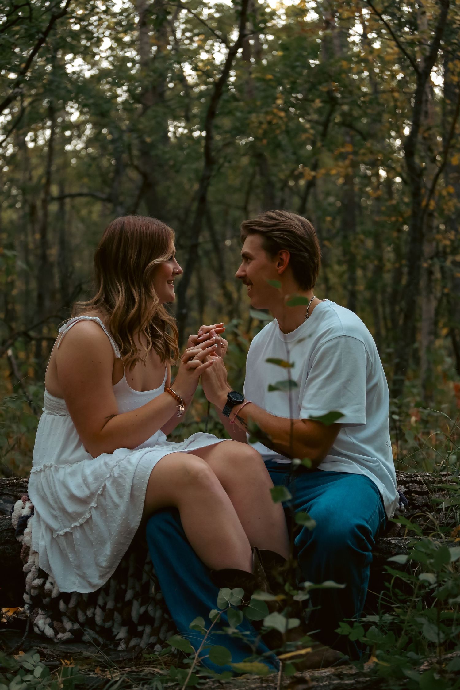 A couple holds hands and smiles at each other while sitting on a log in an autumn forest.