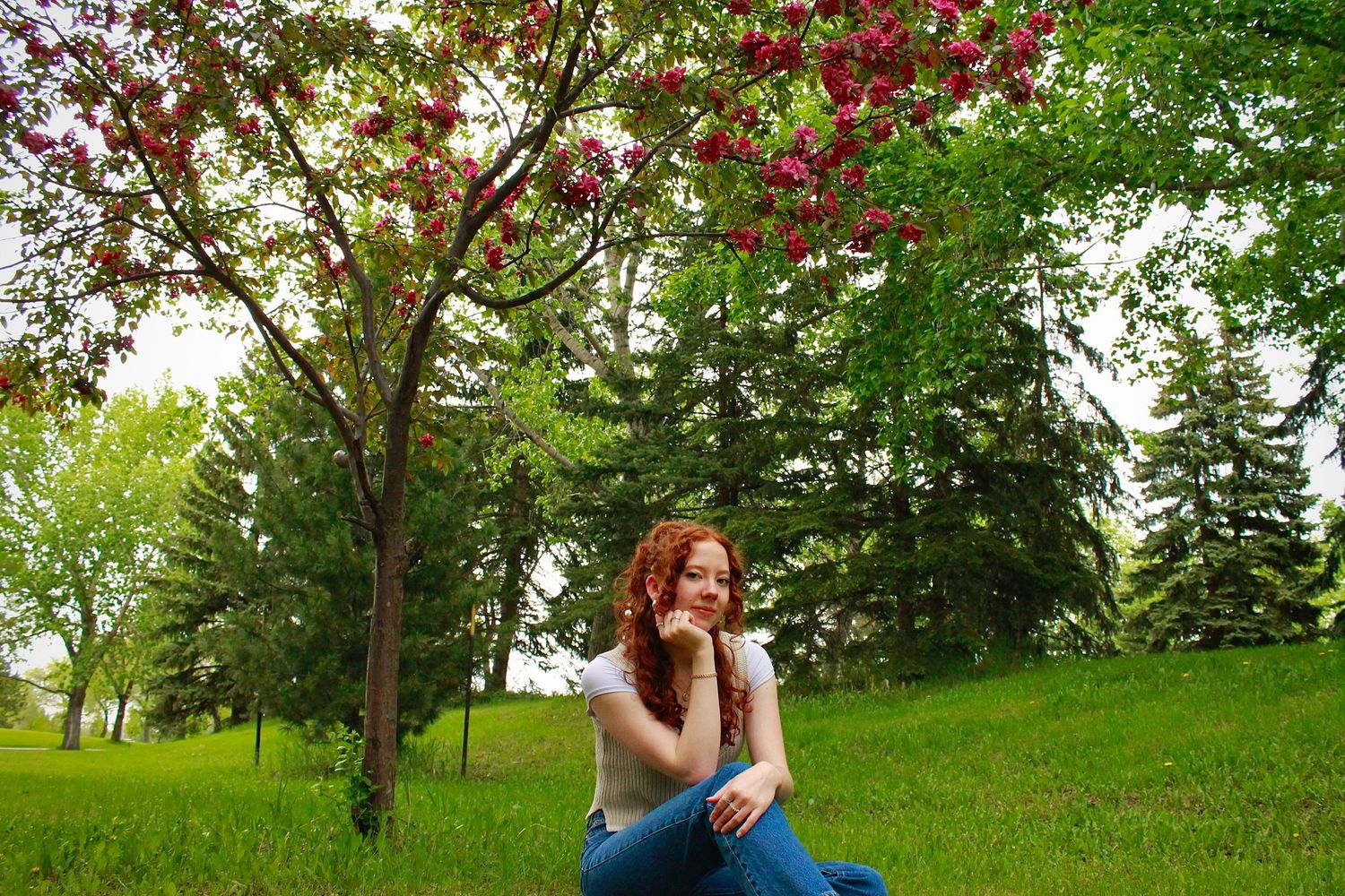 Young redhead woman sitting on grass near a blooming pink flowering tree in a vibrant green park.