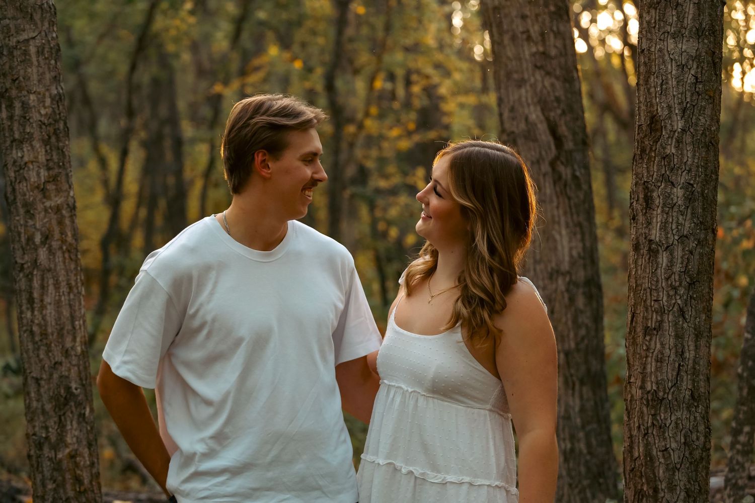 Young couple in white outfits smiling at each other in a sunlit autumn forest.