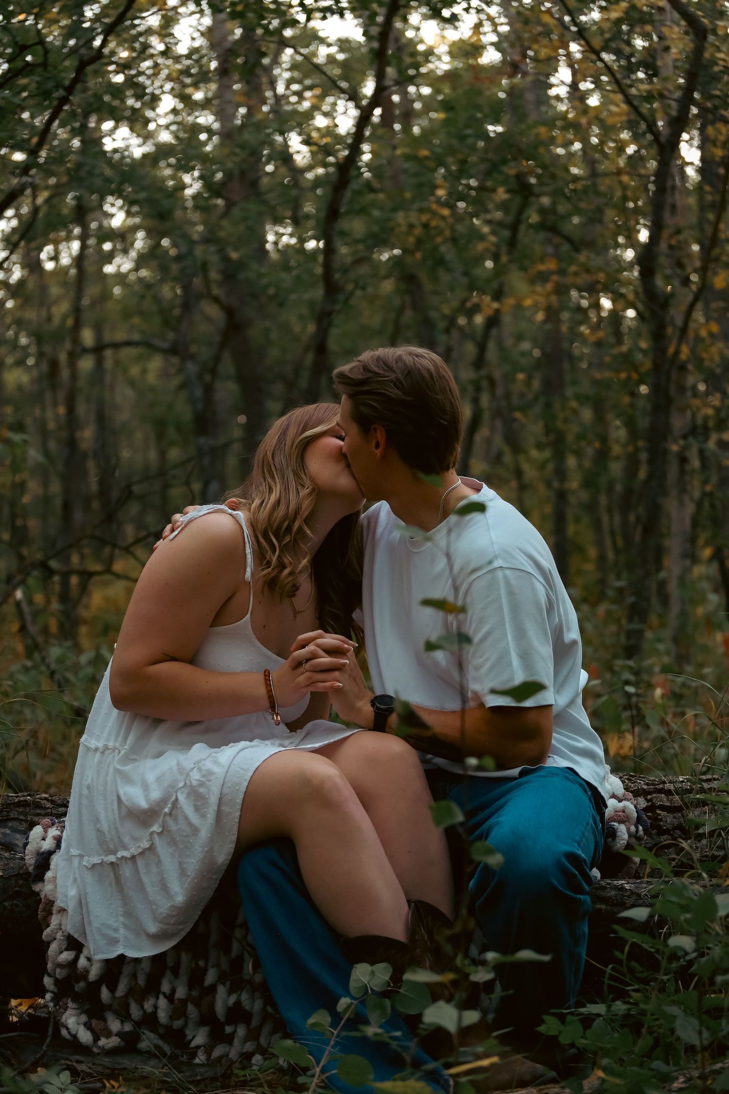 A couple shares a tender kiss while sitting on a log in a lush green forest setting.