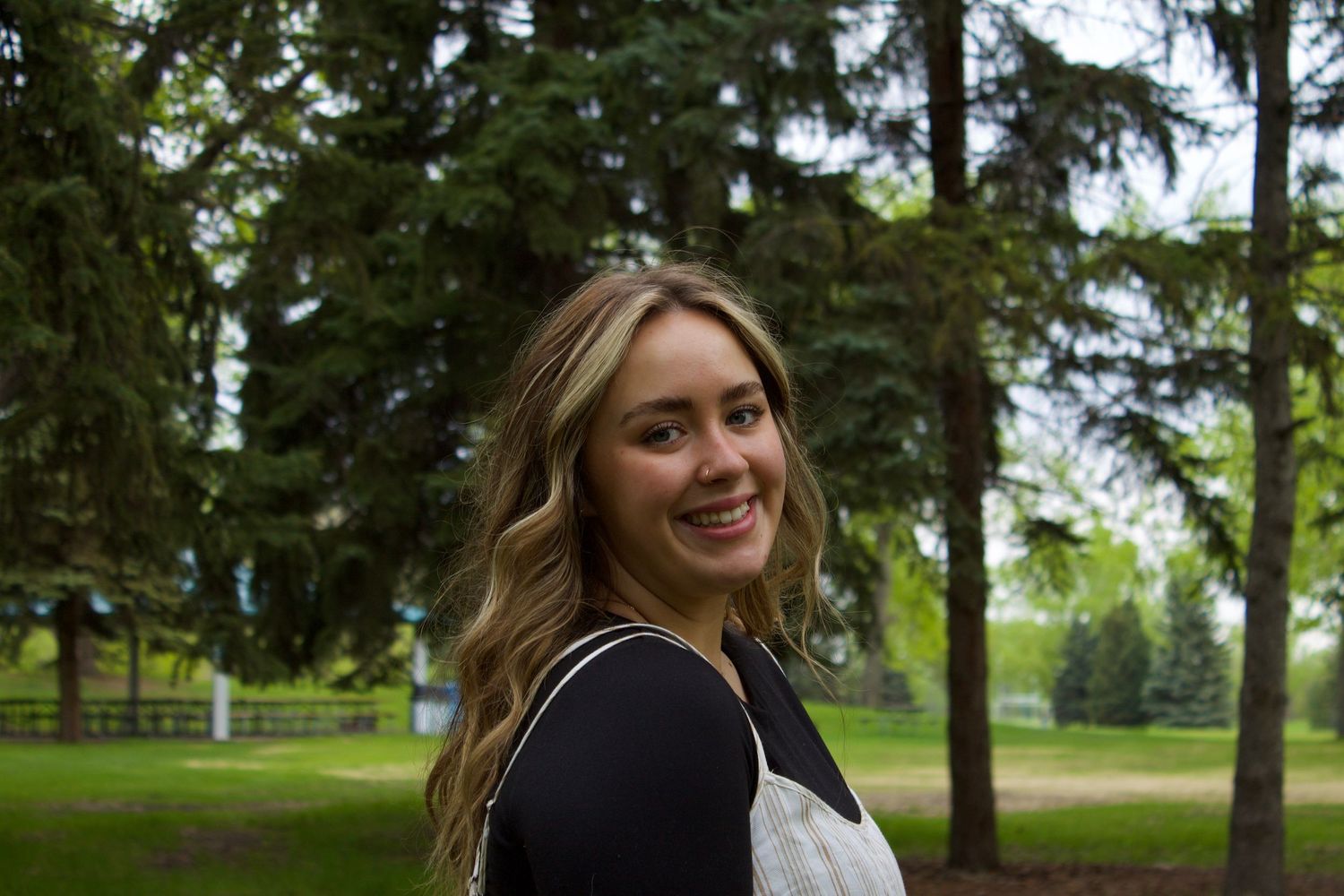 Smiling young woman with wavy blonde hair wearing a black top and overalls in a green park setting.