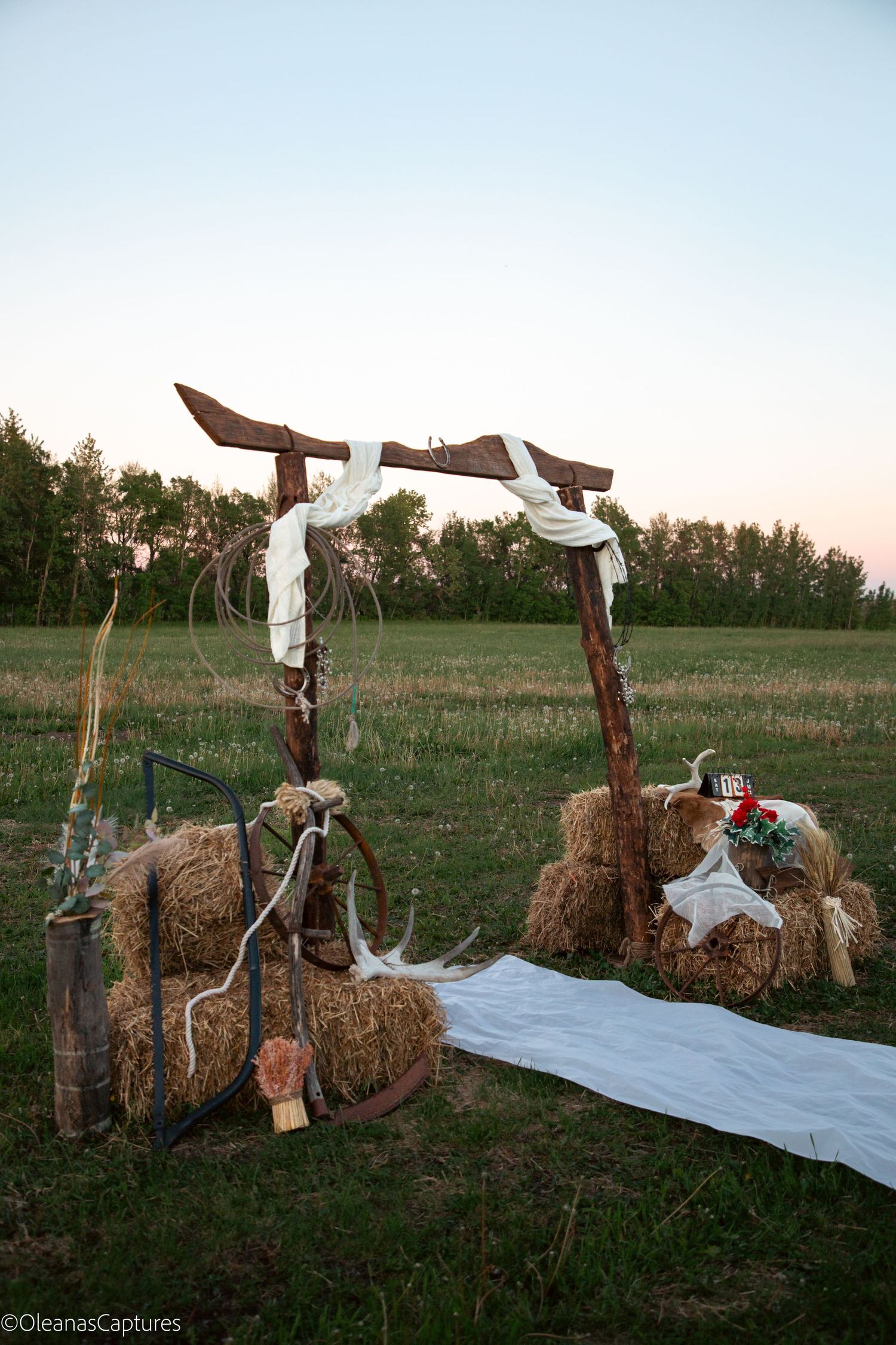 Rustic outdoor wedding ceremony arch with hay bales, white draping, lasso, and red floral accents at dusk.