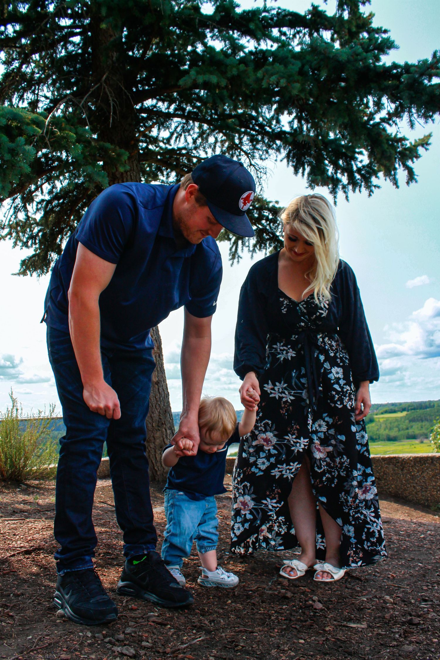 Family of three with toddler learning to walk outdoors under pine tree on sunny day with green landscape.