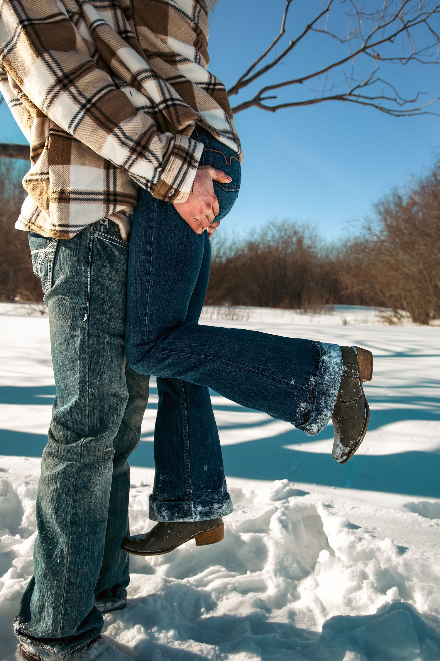 Couple in plaid jackets and jeans embracing closely in a snowy winter landscape outdoors.