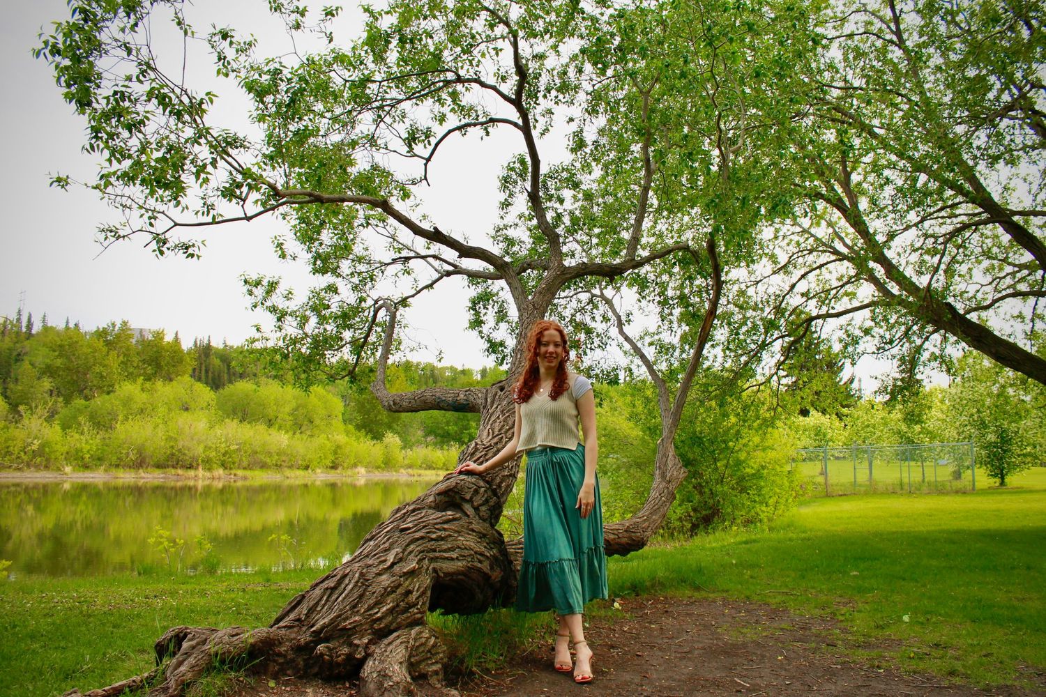 Red-haired woman in teal skirt and beige top leans against a fallen log near a peaceful park lake.