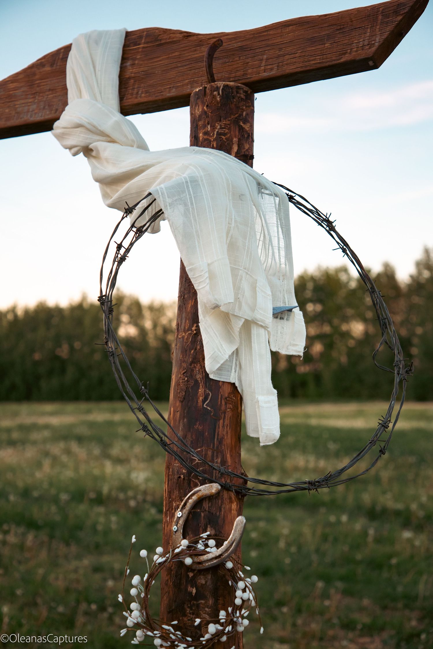 Rustic wooden wedding arch post decorated with barbed wire wreath, horseshoe, and white draped fabric at sunset.