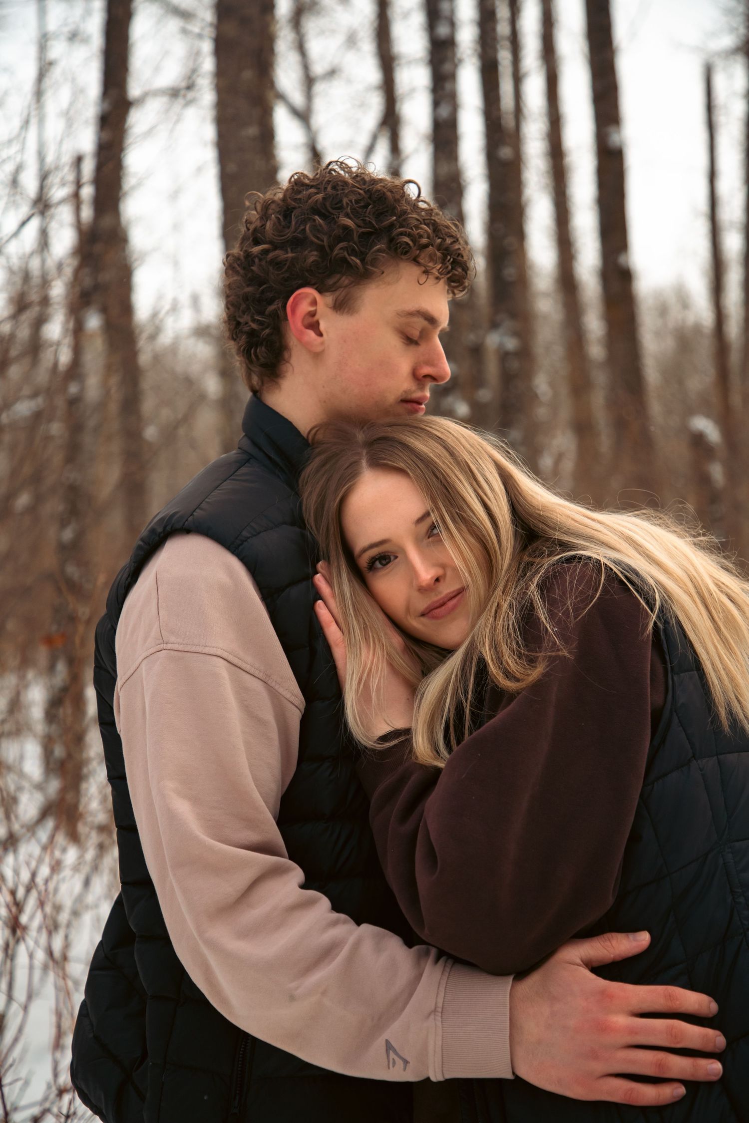 Man tenderly embracing woman from behind as she leans into him during a snowy winter engagement shoot.