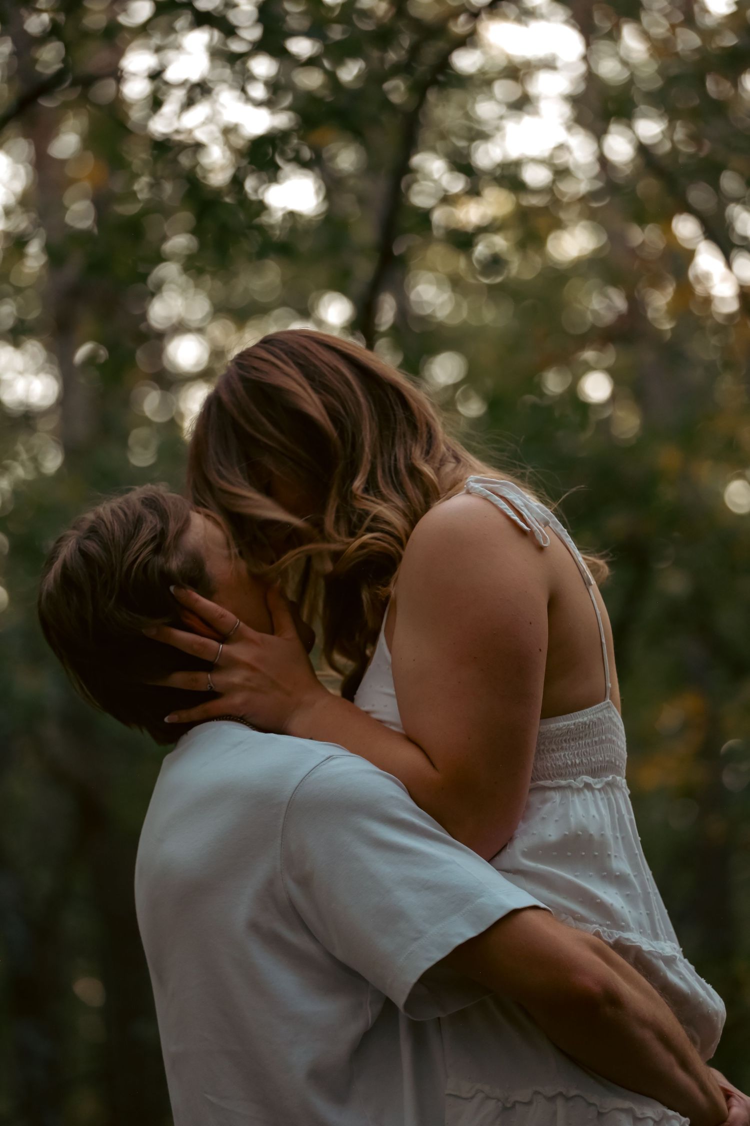 A man holds a woman in a white dress close as she pulls him in for a kiss outdoors.