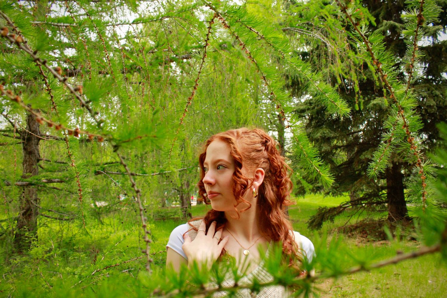 Young redheaded woman gazing thoughtfully among lush green weeping tree branches in a serene park.