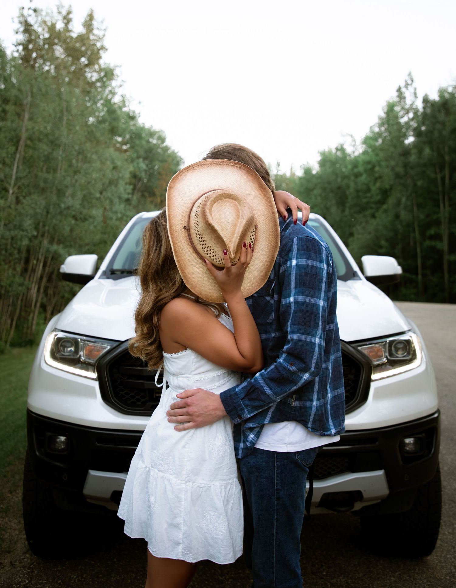 Couple kissing behind a cowboy hat in front of a white truck surrounded by trees.