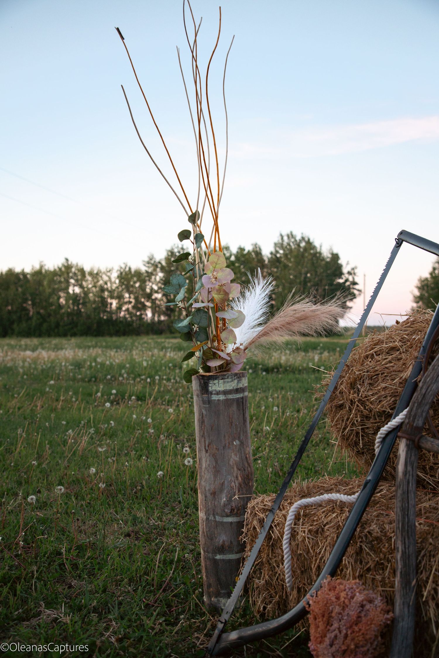 Rustic wooden barrel with dried wildflowers, feathers, and twigs beside a hay bale in an open field at dusk.