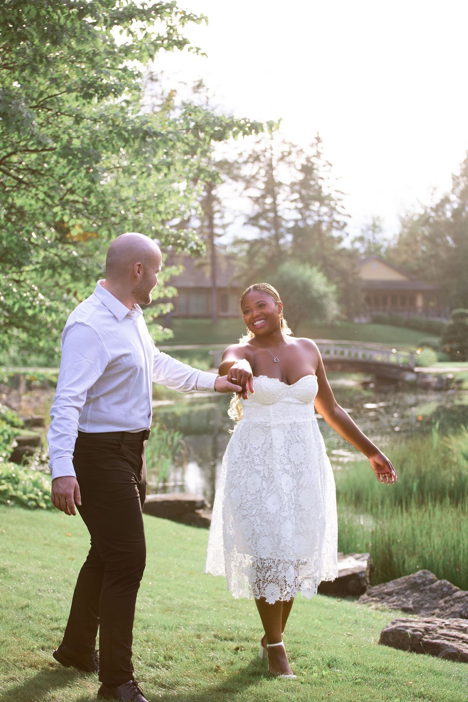 Couple holding hands and walking through a sunlit garden with a pond and bridge in the background.
