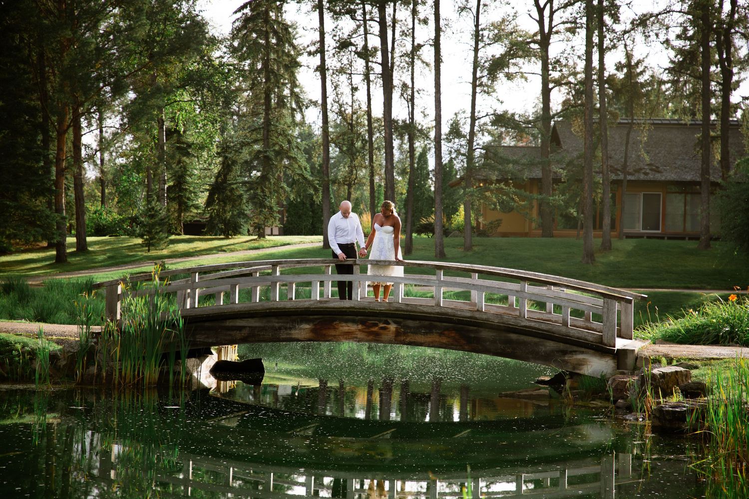 Bride and groom standing together on a wooden arched bridge reflected in a serene garden pond.