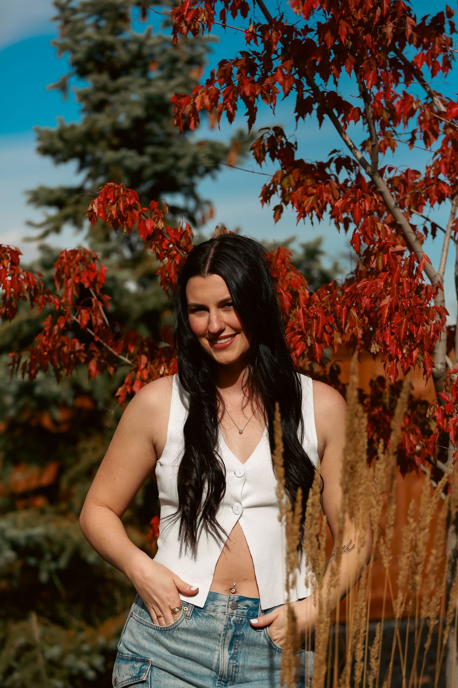 Smiling brunette woman in white crop top and jeans posing outdoors with vibrant red autumn foliage background.