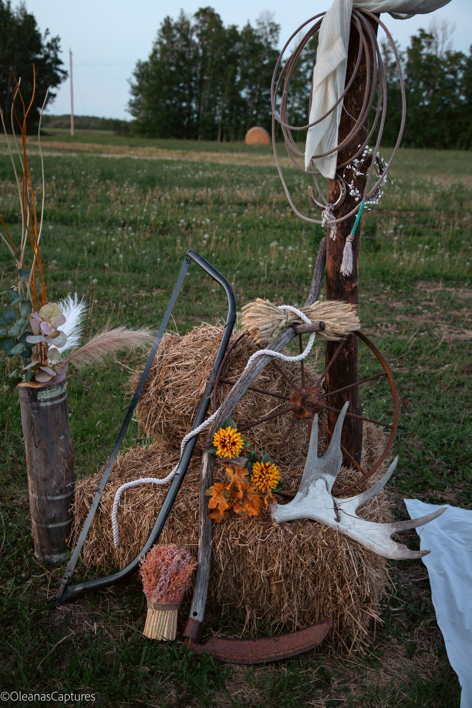 Rustic outdoor wedding arch decor with hay bales, deer antlers, sunflowers, farm tools, and dream catchers in a field.