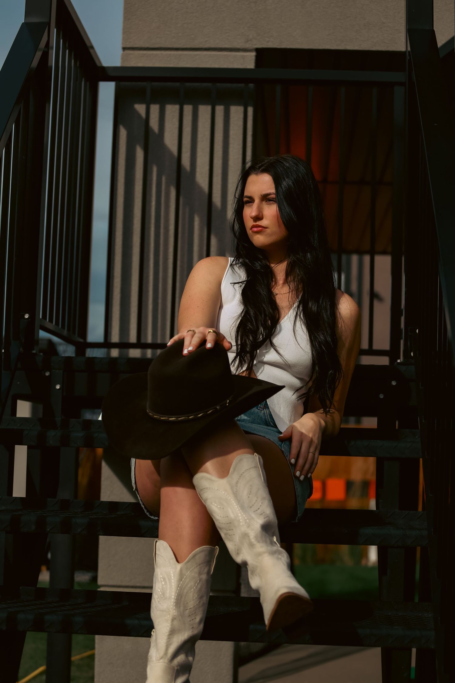 Brunette woman in white top, denim skirt, and white cowboy boots poses on metal stairs with black hat.